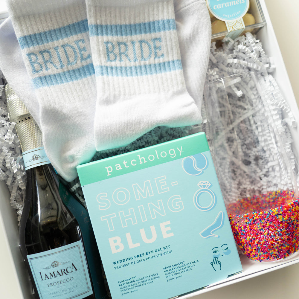 Photograph of a bridal gift box featuring a Patchology eye gel kit, light blue striped BRIDE socks, and a bottle of La Marca Prosecco next to a glass filled with vibrant rainbow sprinkles