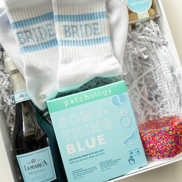Photograph of a bridal gift box featuring a Patchology eye gel kit, light blue striped BRIDE socks, and a bottle of La Marca Prosecco next to a glass filled with vibrant rainbow sprinkles
