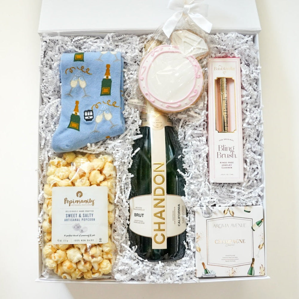 Photograph of a curated engagement gift box featuring a bottle of Chandon Brut sparkling wine surrounded by a pink-rimmed engagement ring sugar cookie, light blue champagne-patterned socks, and a bag of Popinsanity popcorn on white shredded paper