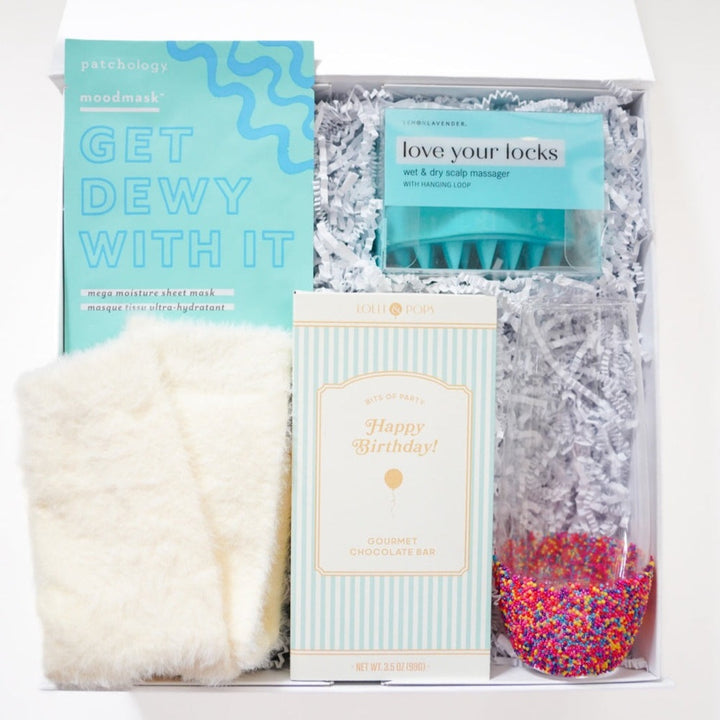 Studio photograph of a curated birthday gift box featuring a Patchology sheet mask, a Lolli &amp; Pops chocolate bar, a teal scalp massager, and a glass flute with a vibrant rainbow sprinkle base nestled in white shredded paper
