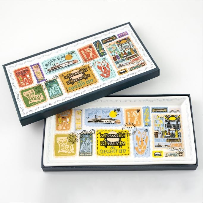 Ceramic tray and illustrated box by Rifle Paper Co. featuring New Orleans landmarks such as the Crescent City Bridge and St. Louis Cathedral, accented with a vibrant red crawfish, a yellow Mississippi River steamboat, and colorful vintage-style postage stamps