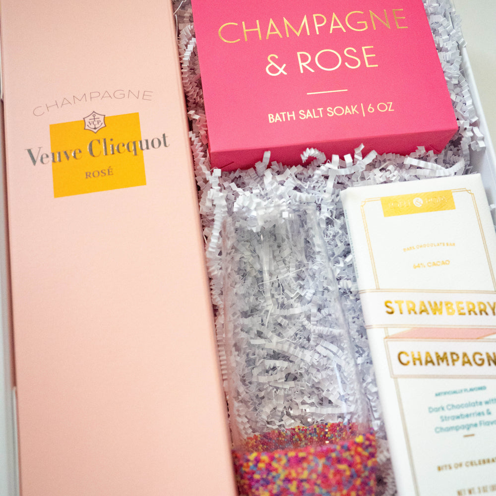 Photograph of a pink-themed luxury gift box featuring a Veuve Clicquot Rosé Champagne box, a bright pink bath salt soak with gold foil lettering, and a glass flute filled with vibrant multi-colored sprinkles resting on white crinkle paper filler