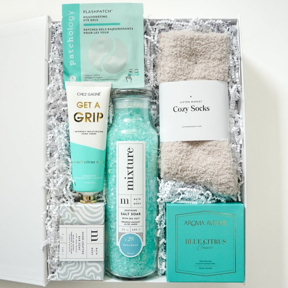 Photograph of a Giften Market wellness gift box containing turquoise Mixture bath salts in a tall glass bottle, fuzzy beige socks, and a mint green Aroma Avenue candle box on white crinkle paper