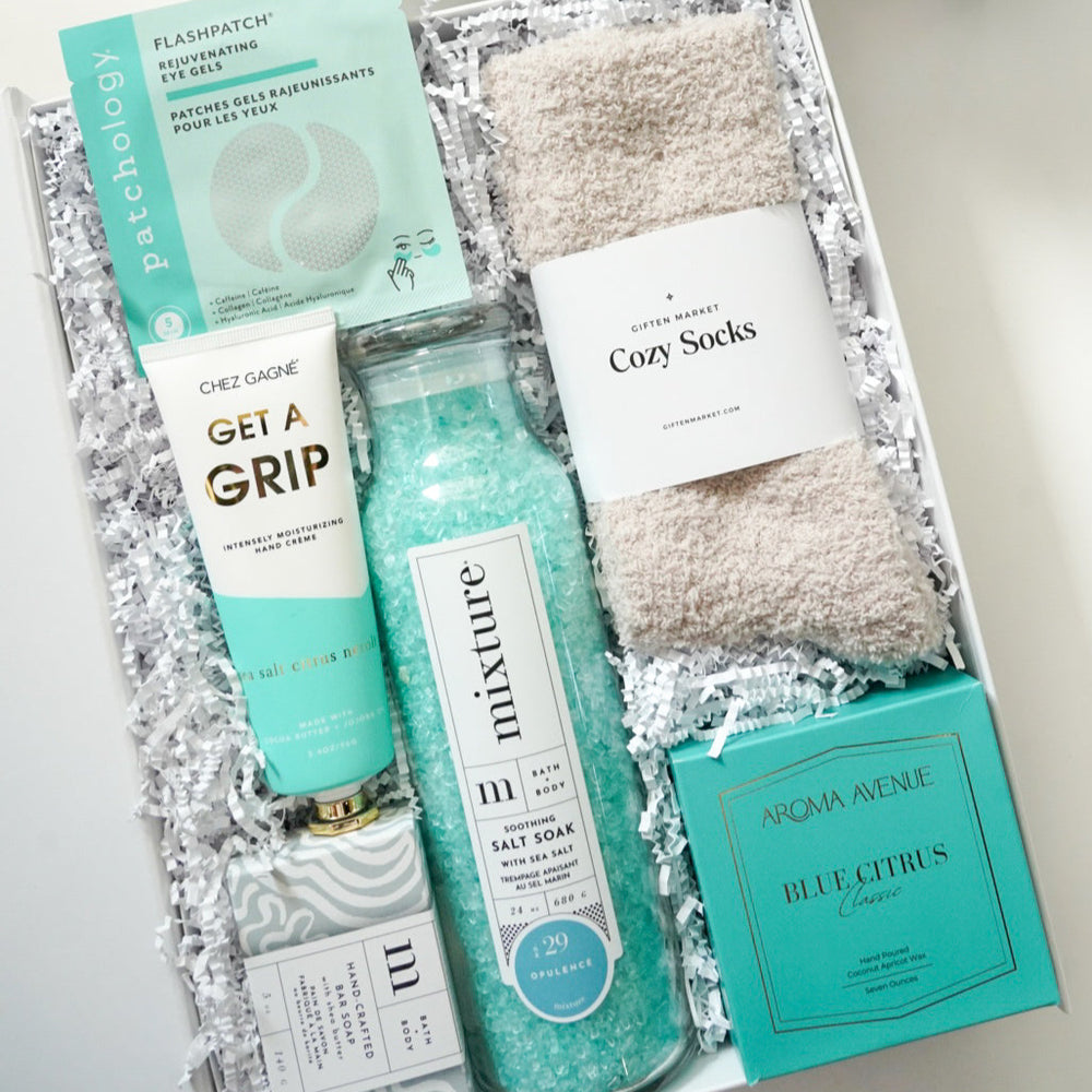 Digital photograph by Giften Market of a curated self-care gift box featuring Patchology eye gels and an Aroma Avenue candle alongside vibrant teal sea salts and fluffy beige cozy socks nestled in white crinkle paper