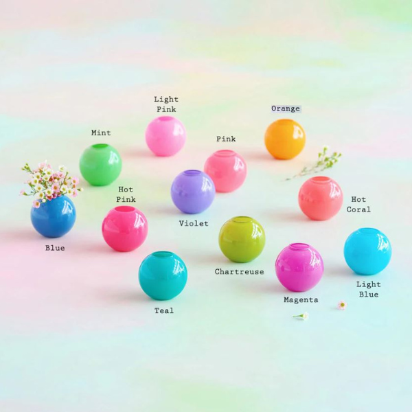 Studio photography of a collection of small glossy round bud vases arranged on a soft pastel gradient background featuring color labels like Teal and Chartreuse and a single blue vase holding a tiny bouquet of pink and white wildflowers