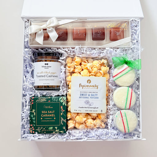 An open white gift box filled with assorted gourmet treats on a white background. The box contains small chocolate bars, a jar of salted cashews, a bag of sea salt caramels, a bag of peppermint candy corn, a white and caramel cookie, and macarons, all arranged in white crinkle paper.