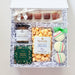 An open white gift box filled with assorted gourmet treats on a white background. The box contains small chocolate bars, a jar of salted cashews, a bag of sea salt caramels, a bag of peppermint candy corn, a white and caramel cookie, and macarons, all arranged in white crinkle paper.