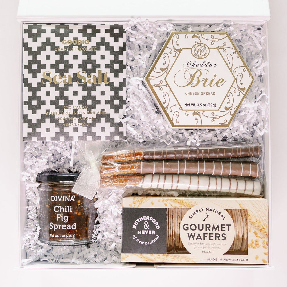 Studio photograph of a luxury gourmet gift box featuring Goodio Sea Salt chocolate, Cheddar Brie cheese spread, and Divina Chili Fig spread nestled in white crinkle paper with striped chocolate-dipped pretzel rods and Rutherford & Meyer wafers