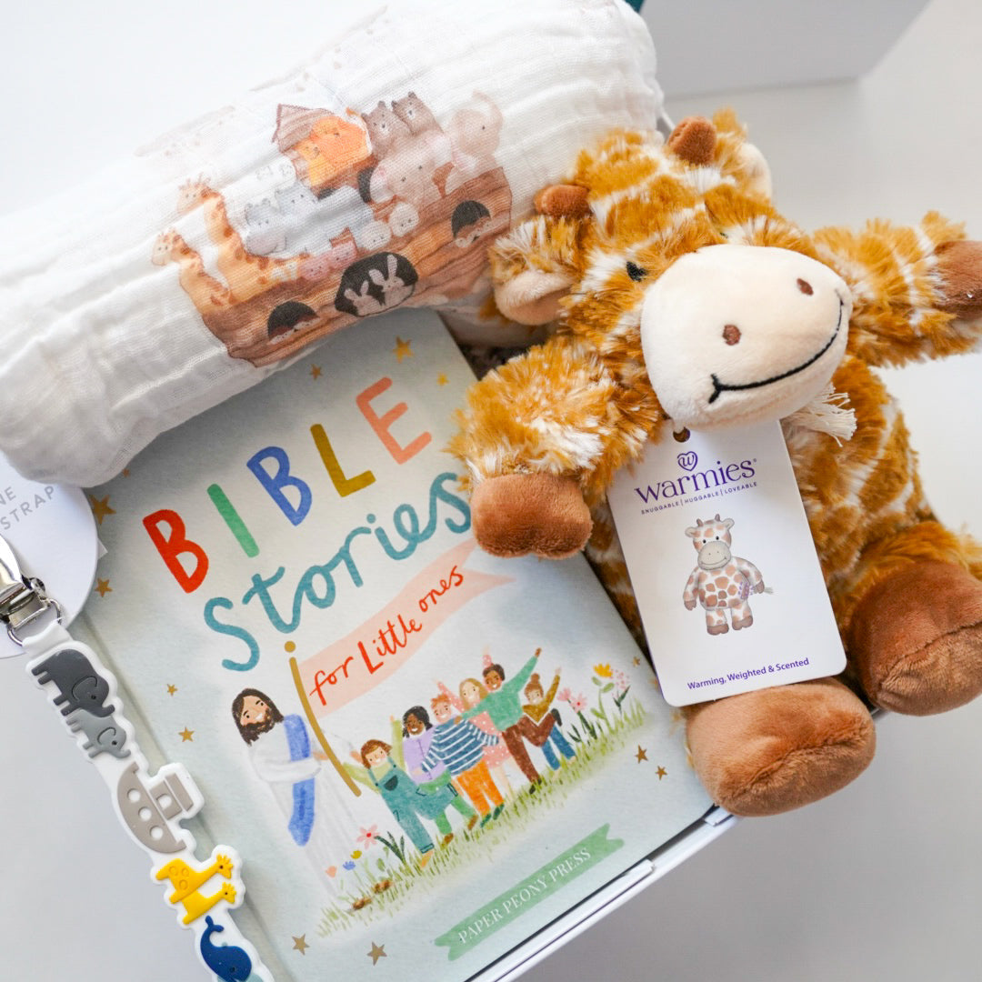 Photograph of a baby gift set by Paper Peony Press and Warmies featuring the Bible Stories for Little Ones book with rainbow lettering, a plush giraffe, a Noah&