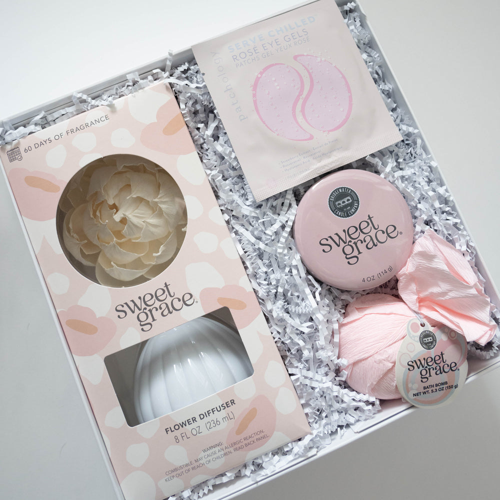 Photograph of a Bridgewater Candle Company self-care gift box featuring Sweet Grace branded items including a cream-colored paper flower diffuser, a pink-wrapped bath bomb, and a packet of Patchology Rosé Eye Gels nestled in white crinkle-cut filler paper