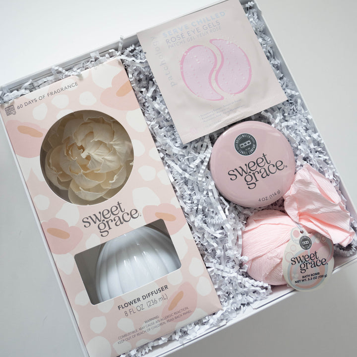 Photograph of a Bridgewater Candle Company self-care gift box featuring Sweet Grace branded items including a cream-colored paper flower diffuser, a pink-wrapped bath bomb, and a packet of Patchology Rosé Eye Gels nestled in white crinkle-cut filler paper