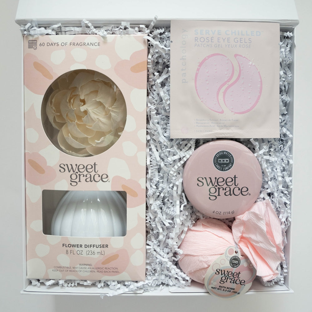 Product photography of a Sweet Grace and Patchology beauty gift set in a white box featuring a cream-colored flower diffuser, pink Rosé eye gels, and a light pink paper-wrapped bath bomb on white crinkle paper