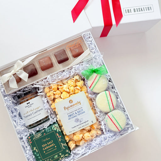 An open gift box filled with assorted treats and packaged snacks, shown next to its white lid tied with a red ribbon. The contents include chocolates, macarons, a small jar, and a green carded item on a light neutral background.