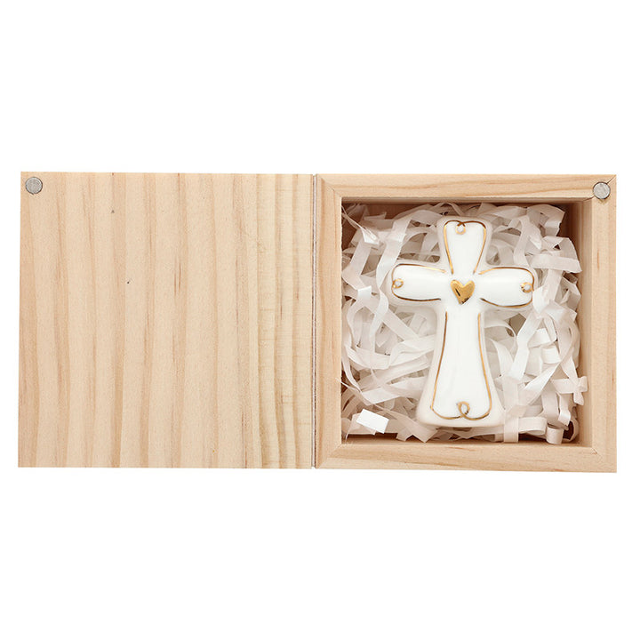 White ceramic cross featuring a central gold heart and metallic gold trim nestled inside a light-toned wooden gift box on a bed of white shredded paper filler