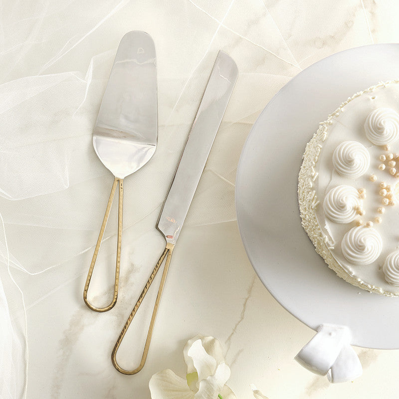 Commercial photography of a stainless steel cake server and knife set with textured gold looped handles positioned on a white marble surface next to a white frosted cake featuring whipped cream swirls and decorative edible pearls