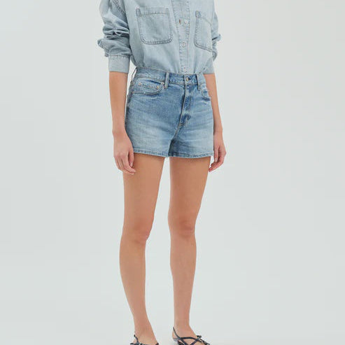 Digital fashion photograph showcasing a model in light-wash high-waisted denim shorts and a matching button-down shirt with chest pockets in a minimalist white studio setting paired with black strappy sandals