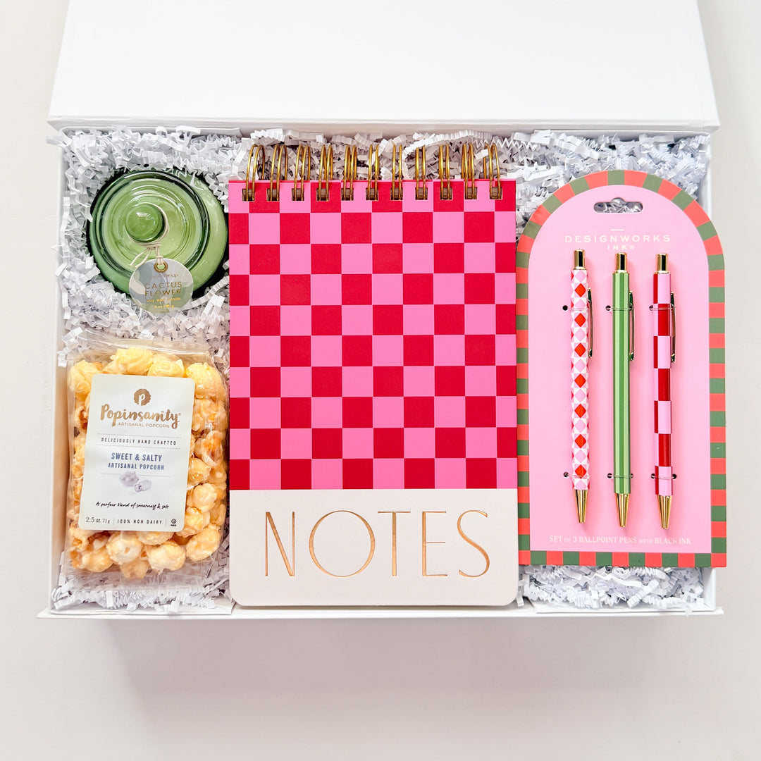 Gift set with a pink checkered notebook, pen, and snacks in a clear box on a white background