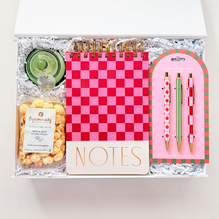 Gift set with a pink checkered notebook, pen, and snacks in a clear box on a white background