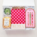 Gift set with a pink checkered notebook, pen, and snacks in a clear box on a white background