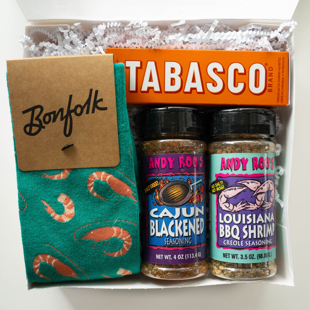 Product photograph of a Louisiana culinary gift box featuring teal Bonfolk socks with an orange shrimp pattern, a bright orange Tabasco brand box, and Andy Roo’s Creole seasoning jars set in white shredded paper