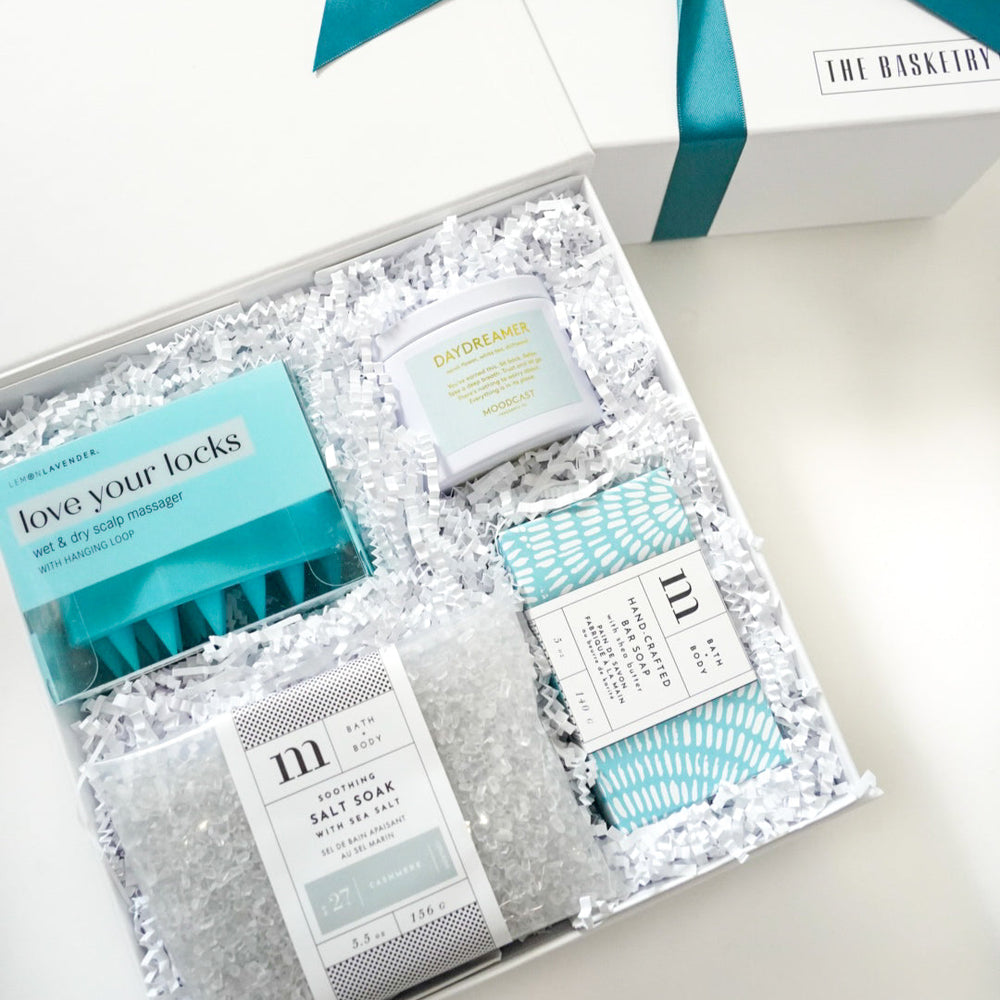 Photograph by The Basketry of a white spa gift box on a bright surface featuring a teal scalp massager, a Daydreamer candle, and soothing bath salts nestled in white crinkle paper with a matching blue ribbon