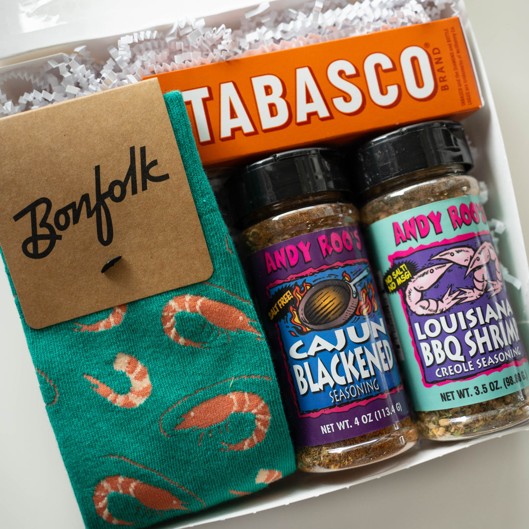 Photograph of a Louisiana-themed gift box featuring teal Bonfolk socks with an orange shrimp pattern, a bright orange Tabasco box, and Andy Roo’s seasoning jars with colorful labels nestled in white shredded paper packing