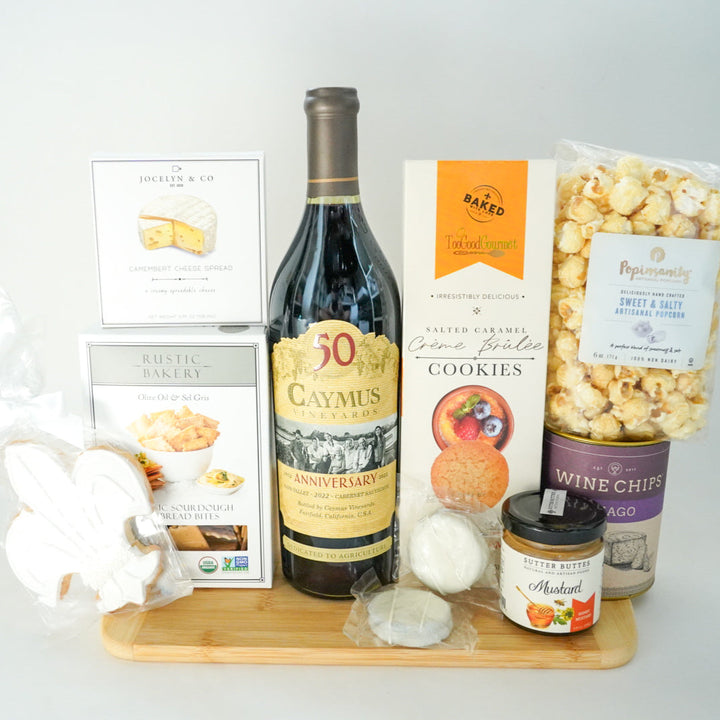 Photograph of a gourmet gift set on a wooden board featuring a Caymus Vineyards 50th Anniversary Cabernet Sauvignon bottle, white-iced fleur-de-lis cookies, and a bag of sweet and salty popcorn