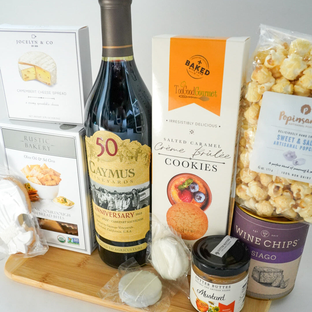 Photograph of a gourmet food gift set on a light wood surface featuring a bottle of Caymus 50th Anniversary Cabernet Sauvignon, vibrant orange Salted Caramel Crème Brûlée cookie packaging, and a purple canister of Asiago Wine Chips