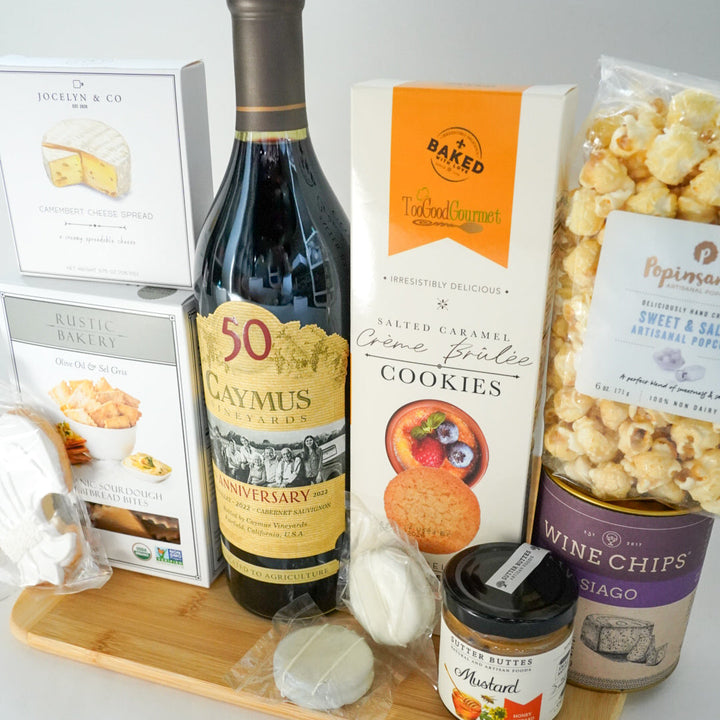 Photograph of a gourmet food gift set on a light wood surface featuring a bottle of Caymus 50th Anniversary Cabernet Sauvignon, vibrant orange Salted Caramel Crème Brûlée cookie packaging, and a purple canister of Asiago Wine Chips
