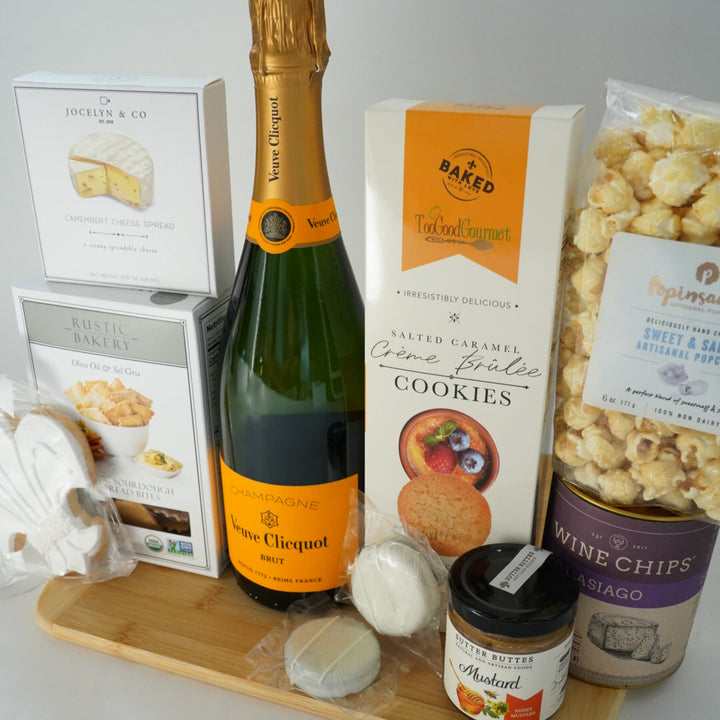Photograph of a luxury gourmet gift collection on a bamboo board featuring a bottle of Veuve Clicquot champagne with its signature orange label, Popinsanity popcorn, and white-wrapped treats in the foreground