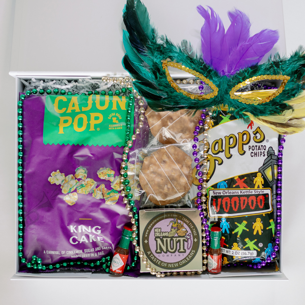 Photograph of a New Orleans Mardi Gras gift box featuring iconic Zapp's Voodoo chips and Cajun Pop popcorn paired with miniature Tabasco bottles, a vibrant green and purple feathered mask, and shimmering metallic beads