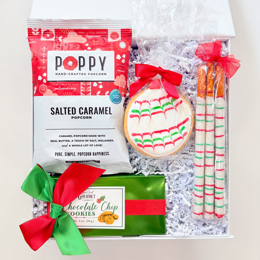 A curated holiday gift box filled with treats, including a bag of Poppy Salted Caramel Popcorn, a decorated cookie with red and green icing, two wrapped candy canes, and a box of chocolate chip cookies, all arranged in white crinkle paper inside an open white box.