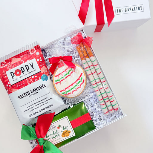 A holiday-themed gift box set from The Basketwork, featuring a white gift box with a red ribbon, a bag of Poppy Salted Caramel cookies, a decorated Christmas cookie, striped candy canes, and a chocolate chip cookie, arranged in white crinkle paper.