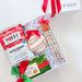 A holiday-themed gift box set from The Basketwork, featuring a white gift box with a red ribbon, a bag of Poppy Salted Caramel cookies, a decorated Christmas cookie, striped candy canes, and a chocolate chip cookie, arranged in white crinkle paper.