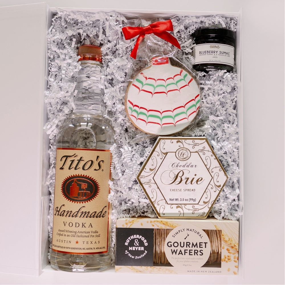 Photograph of a curated holiday gift box featuring Tito's Handmade Vodka from Austin, Texas, alongside gourmet wafers and a festive ornament sugar cookie with a bright red ribbon bow nestled in white crinkled paper filler