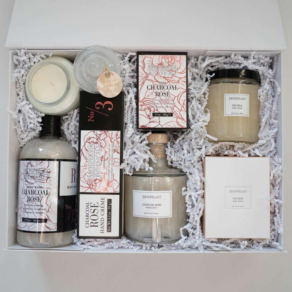 Product photograph by Archipelago Botanicals of a luxury spa gift set in a white box featuring Charcoal Rose and Oat Milk products with detailed red floral illustrations and a glass apothecary-style bottle on white crinkle paper