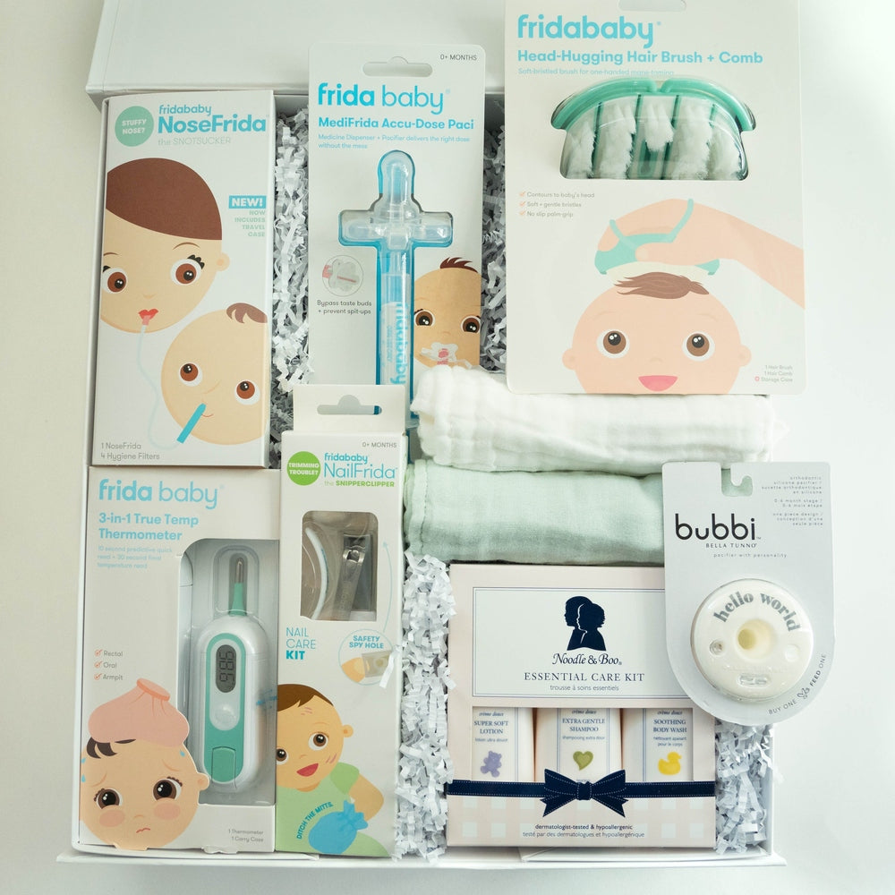 Photograph of a baby gift box featuring Frida Baby health products and a Noodle & Boo care set arranged with mint green washcloths and white shredded paper highlighting whimsical baby face illustrations