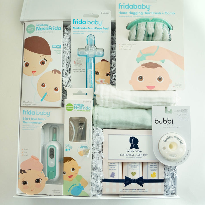 Photograph of a baby gift box featuring Frida Baby health products and a Noodle &amp; Boo care set arranged with mint green washcloths and white shredded paper highlighting whimsical baby face illustrations