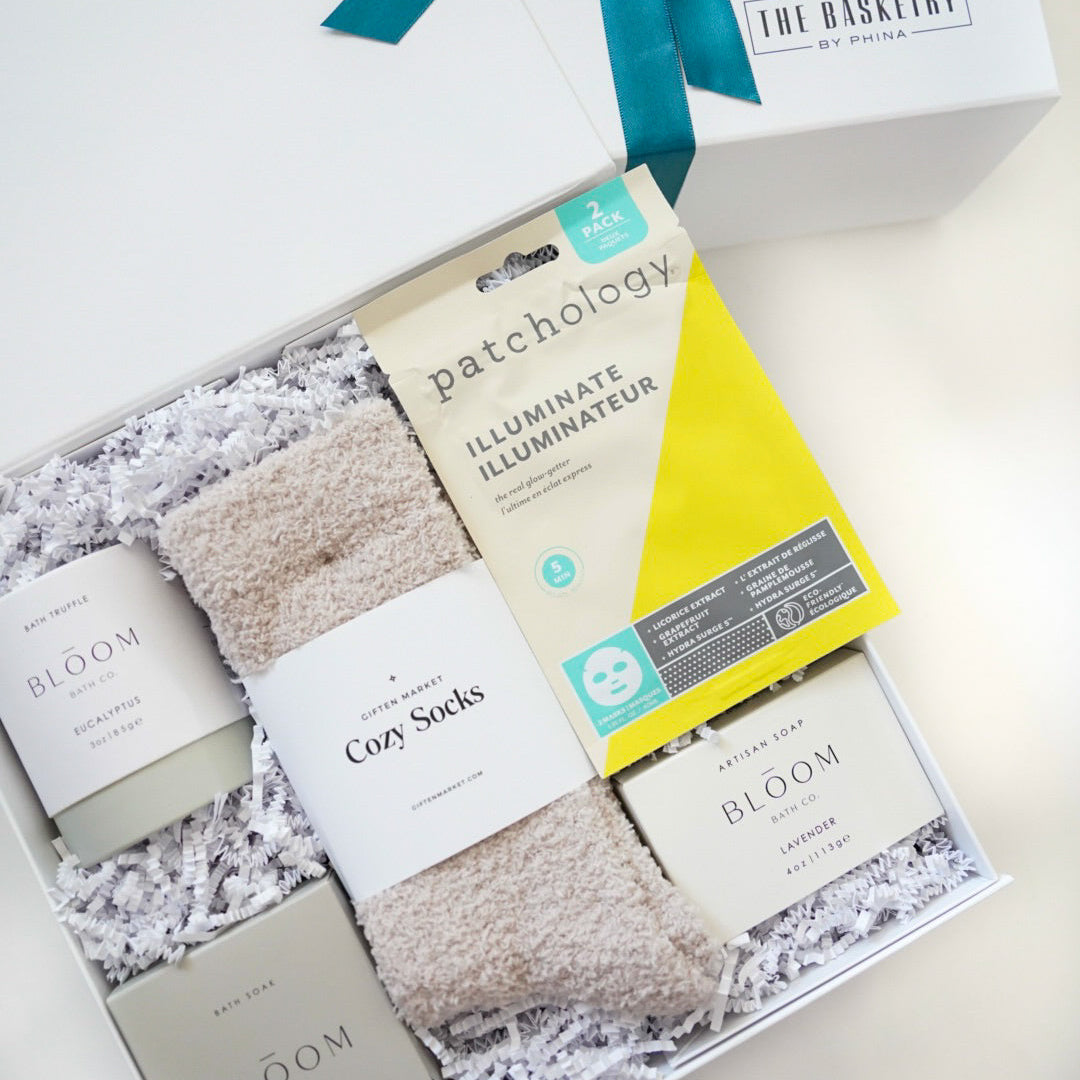 Professional product photograph of a curated self-care gift box by The Basketry by Phina containing vibrant yellow Patchology sheet masks, fuzzy beige cozy socks, and Bloom Bath Co lavender soap nestled in white shredded paper with a teal ribbon accent