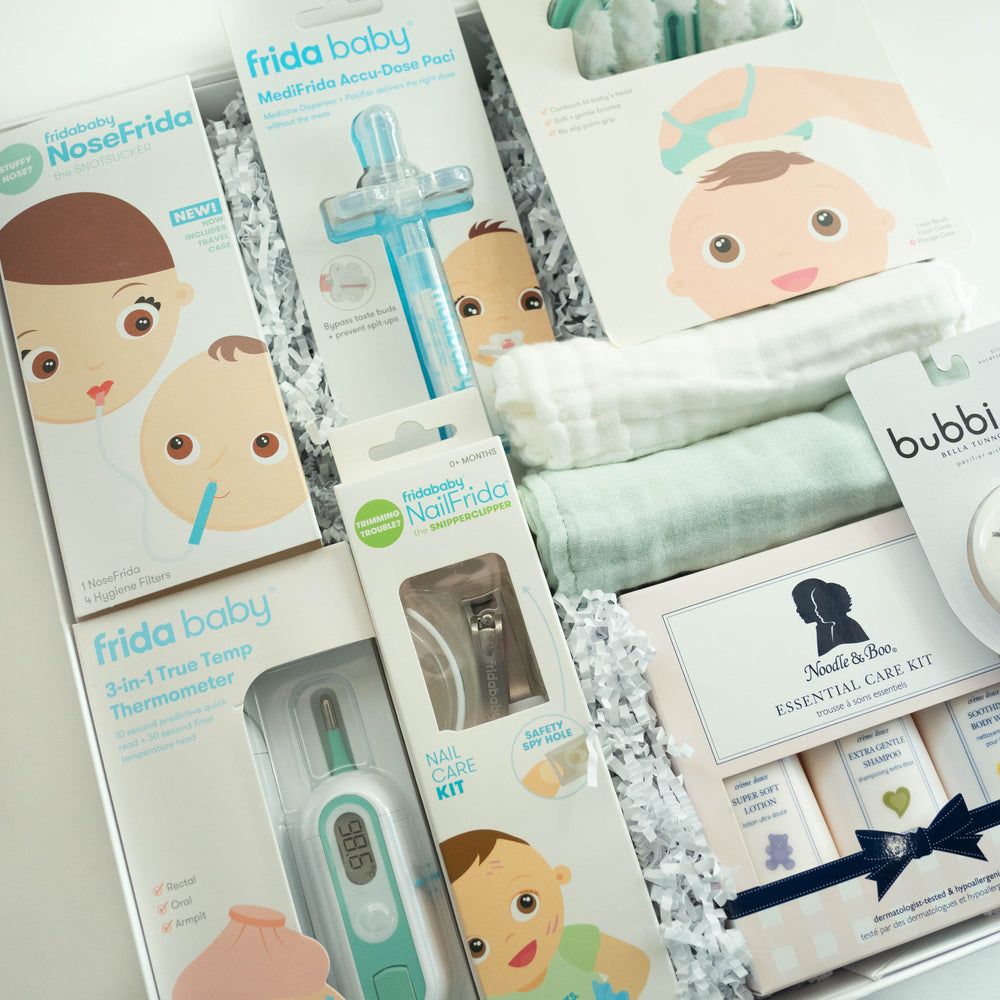 A photograph of a curated baby gift box featuring Frida Baby and Noodle & Boo essentials including a NoseFrida, a digital thermometer displaying 98.6, and a stack of soft mint green and white muslin textiles nestled in white shredded paper
