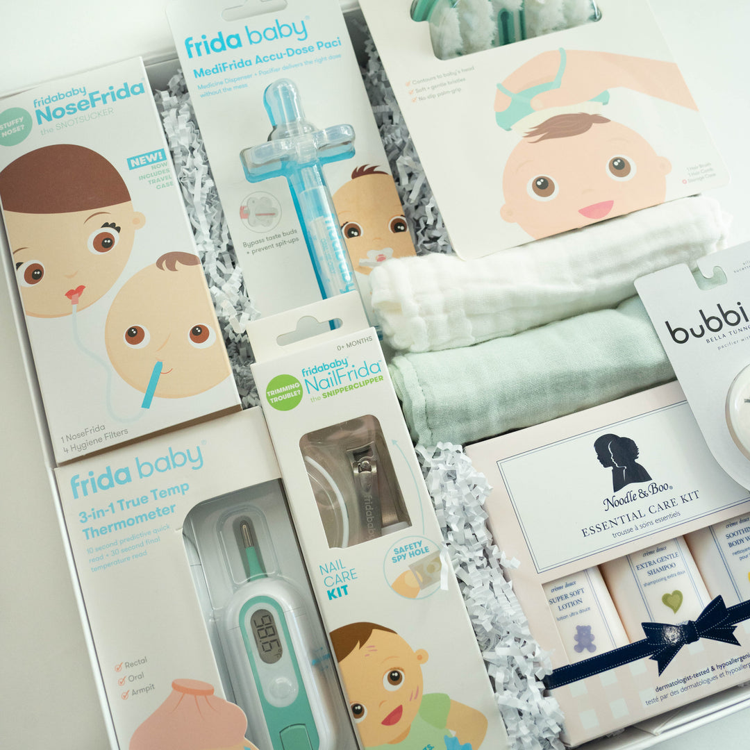 A photograph of a curated baby gift box featuring Frida Baby and Noodle &amp; Boo essentials including a NoseFrida, a digital thermometer displaying 98.6, and a stack of soft mint green and white muslin textiles nestled in white shredded paper