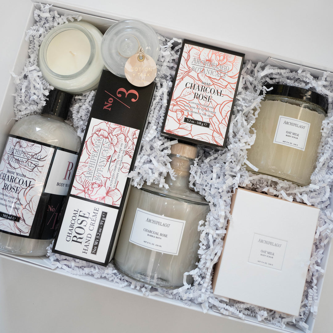 Digital photograph of an Archipelago Botanicals luxury gift set in a white box featuring Charcoal Rose and Oat Milk products nestled in white crinkle paper with pink floral packaging and a cork-stoppered glass bottle