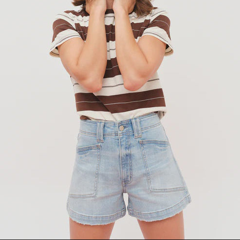 Digital photograph of a model in a neutral studio wearing high-waisted light-wash denim shorts with large front patch pockets and a brown and white horizontal striped t-shirt featuring raw frayed hems