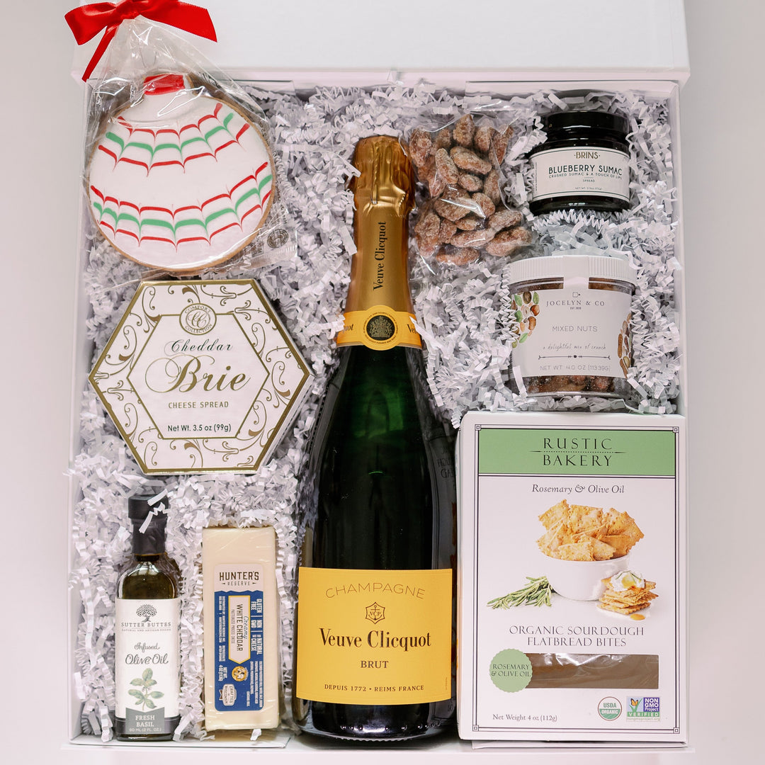 Photograph of a luxury gourmet gift hamper featuring a central bottle of Veuve Clicquot champagne, a festive hand-decorated ornament cookie with red and green icing, and a box of Rustic Bakery flatbread bites nestled in white shredded paper