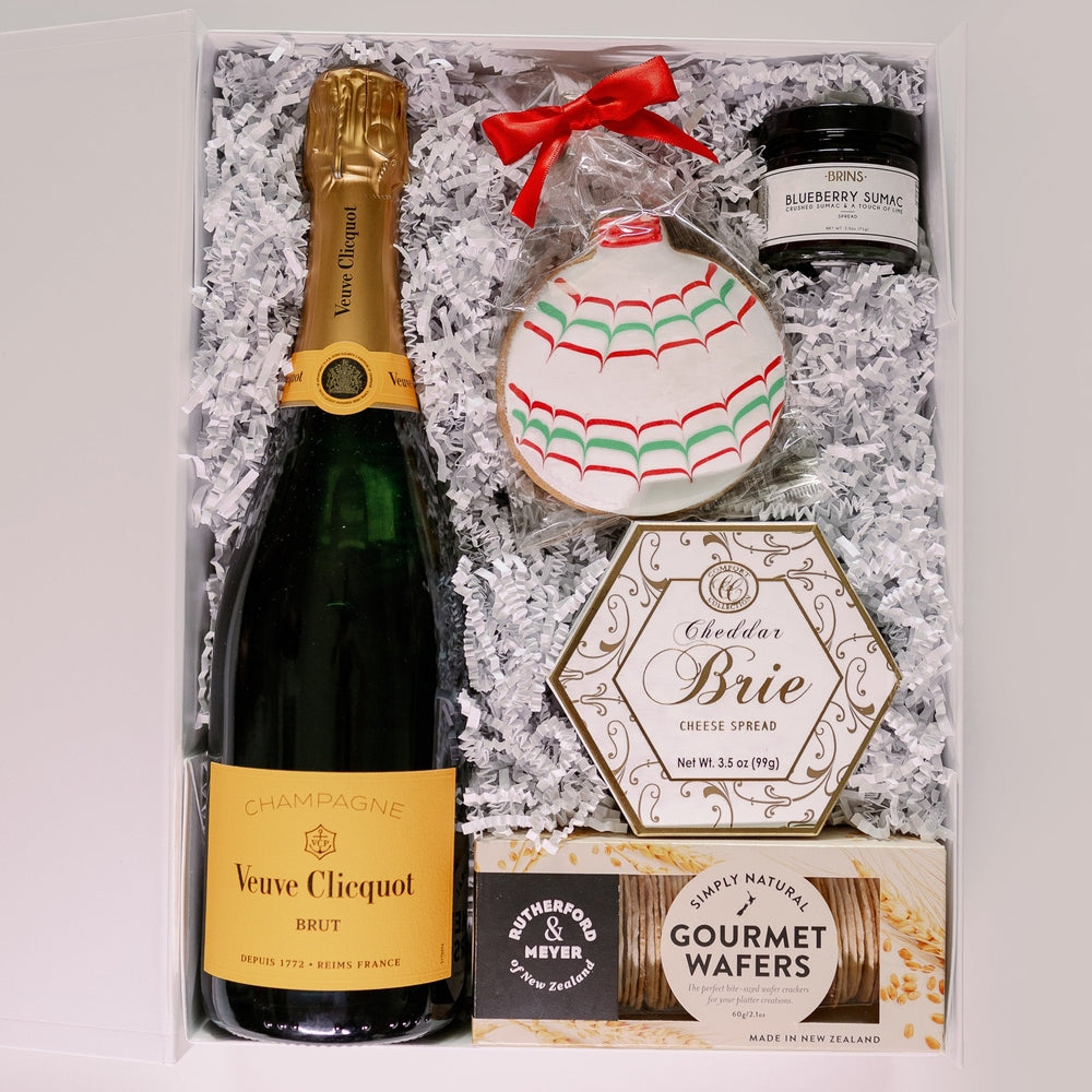 Digital photograph of a luxury holiday gift hamper featuring a bottle of iconic Veuve Clicquot Brut Champagne, a festive ornament-shaped cookie with red and green scalloped icing, and artisanal snacks nestled in white shredded paper padding