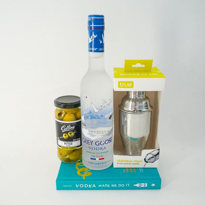 Digital photograph of a cocktail gift set featuring a frosted Grey Goose Vodka bottle with a French Alps label, a jar of green Collins olives, and a boxed stainless steel shaker resting on a vibrant teal book titled Vodka Made Me Do It against a white backdrop