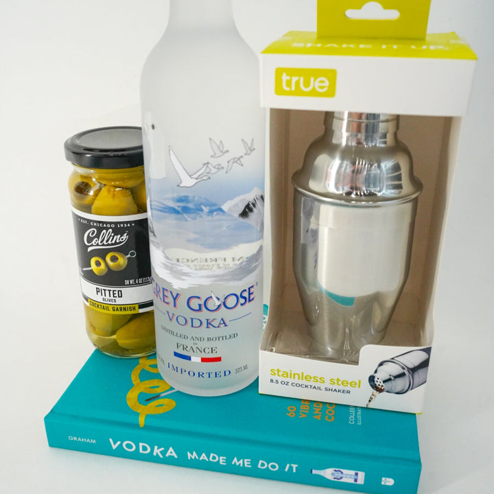 Photograph of a cocktail preparation set featuring a frosted Grey Goose vodka bottle depicting the French Alps, a jar of green olives, and a boxed stainless steel shaker atop a vibrant teal book titled Vodka Made Me Do It against a white background