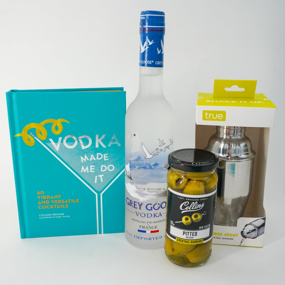 Photograph of a vodka cocktail gift set on a white background featuring the teal book Vodka Made Me Do It by Colleen Graham, a frosted Grey Goose bottle with a blue neck seal, and a stainless steel shaker in a lime yellow box