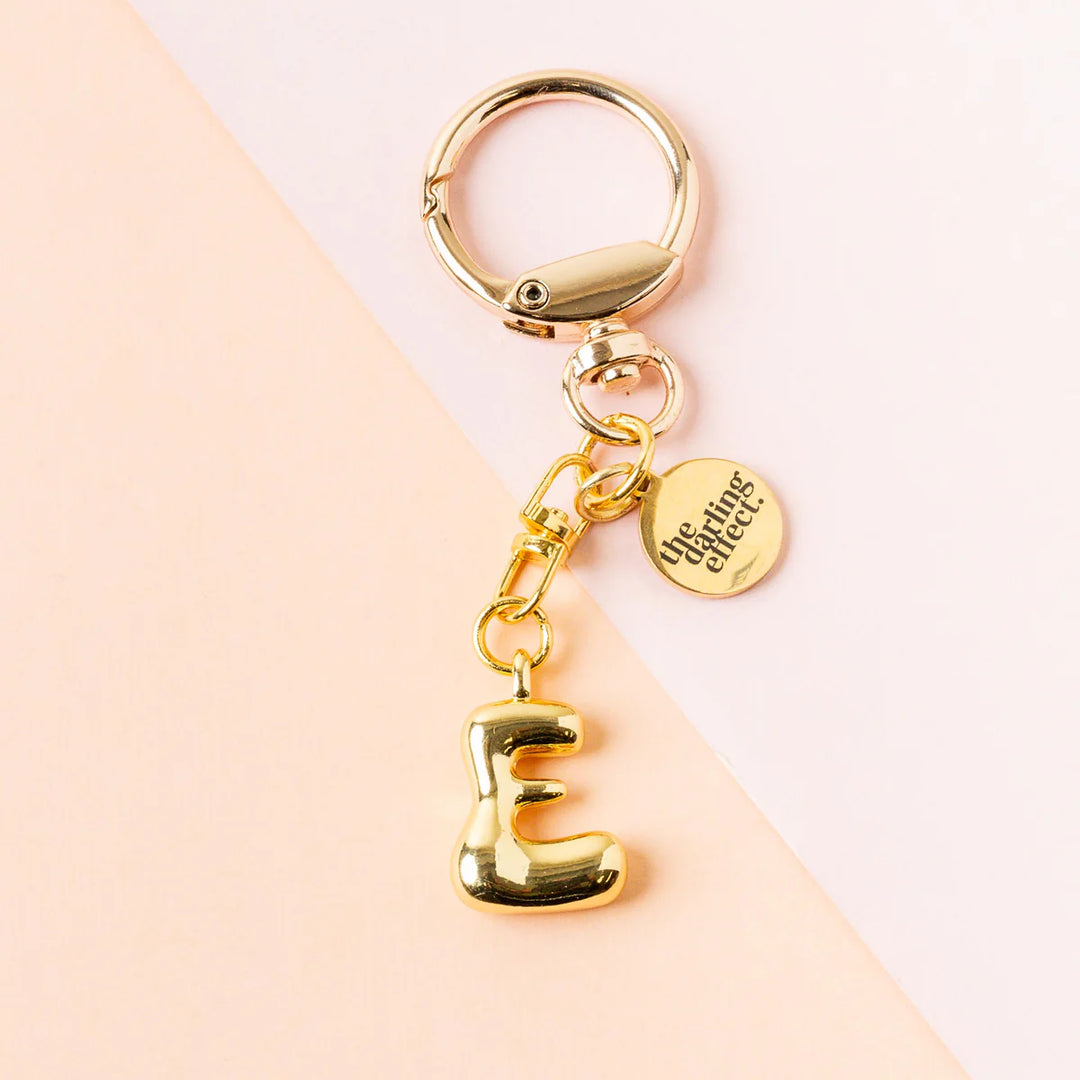 Gold-toned metal keychain by The Darling Effect featuring a puffy balloon-style letter E charm against a two-tone pink background with a circular logo tag and a high-shine lobster claw clasp