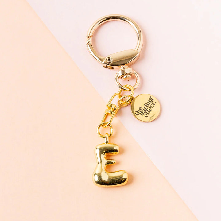 Gold-toned metal keychain by The Darling Effect featuring a puffy balloon-style letter E charm against a two-tone pink background with a circular logo tag and a high-shine lobster claw clasp